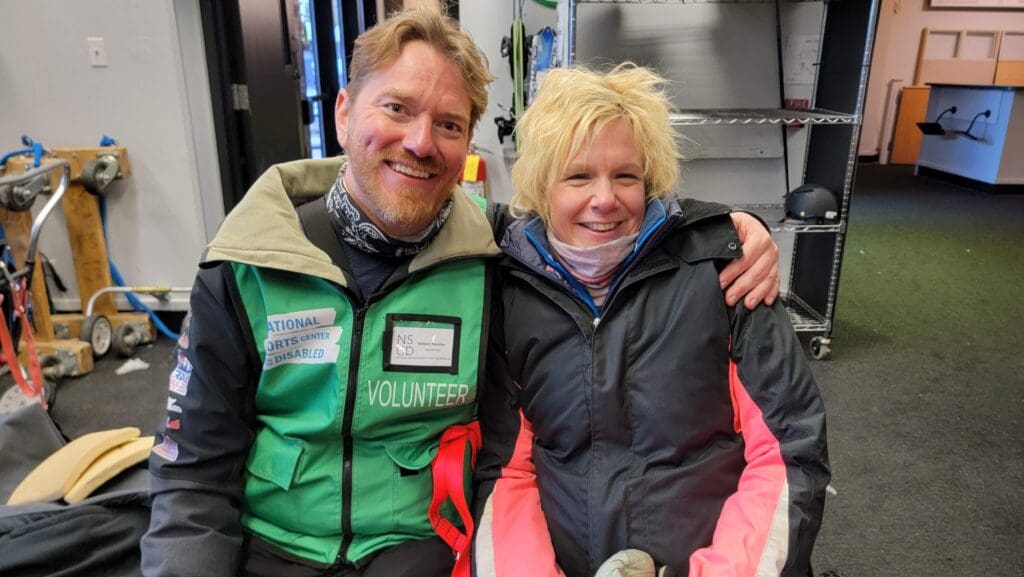 Claire is sitting with her sit-ski instructor in the prep room. They are both smiling