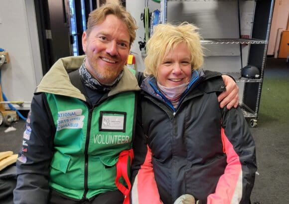 Claire is sitting with her sit-ski instructor in the prep room. They are both smiling