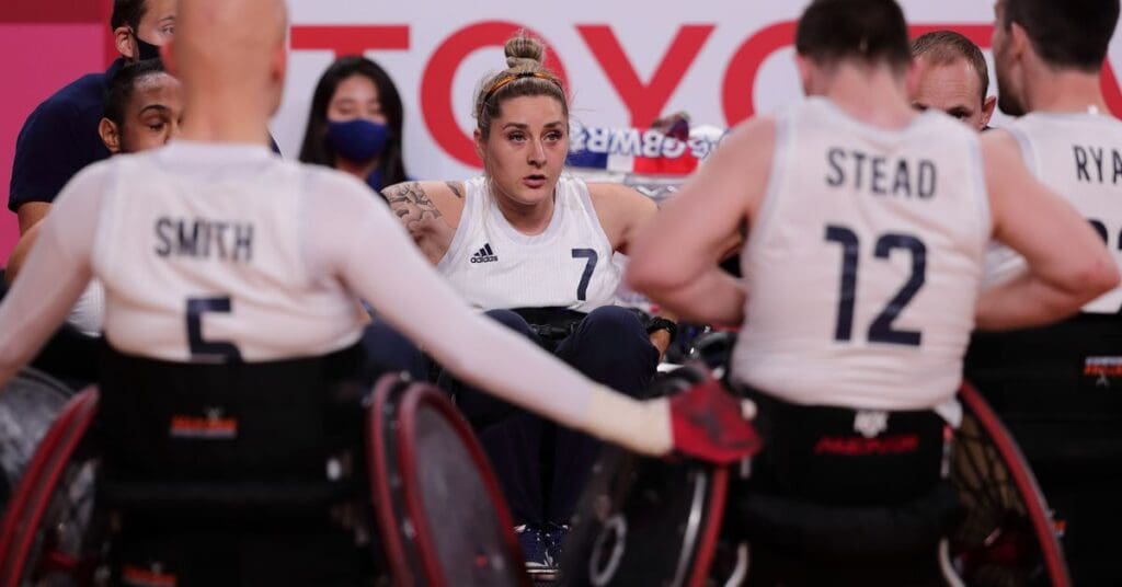 Kylie is wearing her team GB Wheelchair rugby uniform in a huddle with five other players. Kylie has blonde hair in a bun.