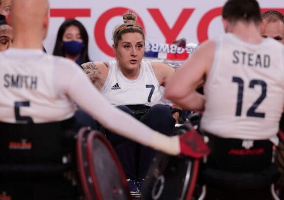 Kylie is wearing her team GB Wheelchair rugby uniform in a huddle with five other players. Kylie has blonde hair in a bun.
