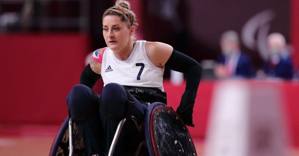 Kylie is wearing her wheelchair rugby uniform for team GB. She is in the middle of a game and has blonde hair tied up in a bun. She looks very focused. 