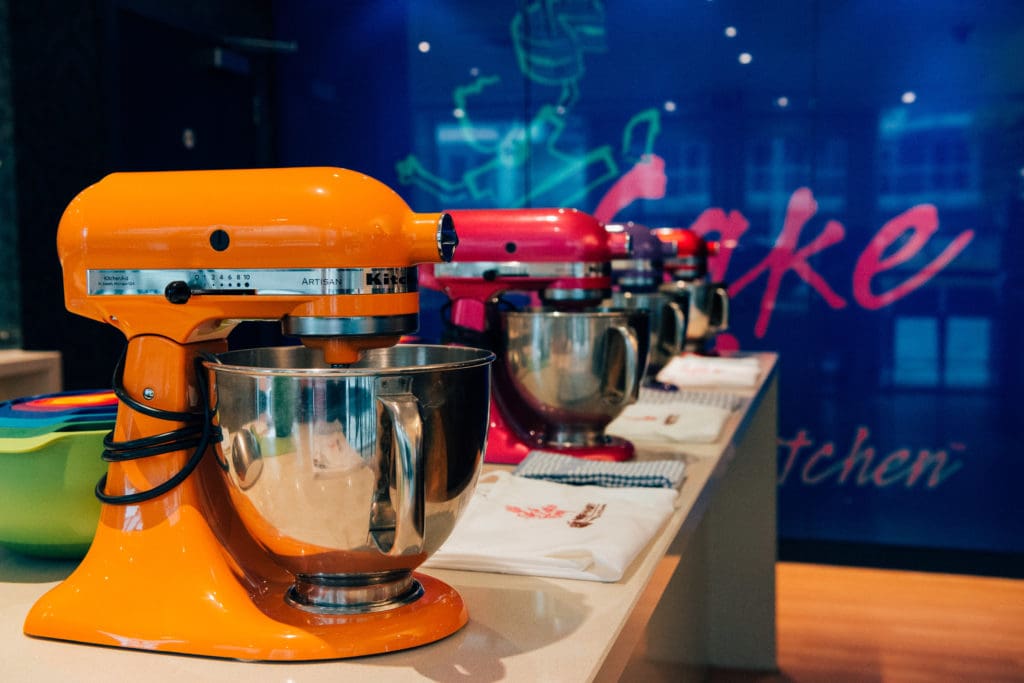 A row of standing mixers for cake making in front of a wall with the Cake Boy logo on
