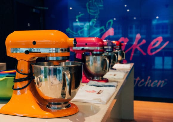 A row of standing mixers for cake making in front of a wall with the Cake Boy logo on