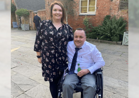 A picture of Karen and her partner who are both smiling on a summers day. Karen accessed our family mentoring service after her partner sustained a spinal cord injury.