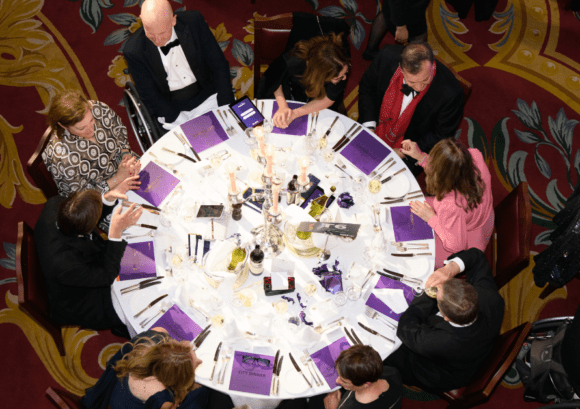 Arial view of one of the tables laid at City Dinner. The table is circular with nine people sat on it and purple brochures at each place setting.