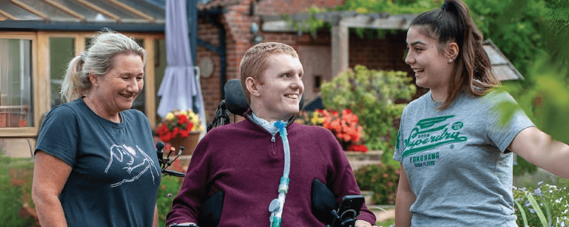 Rob is laughing with his two personal assistants. Rob is wearing a burgundy jumper. They are outside with lots of flowers surrounding them.
