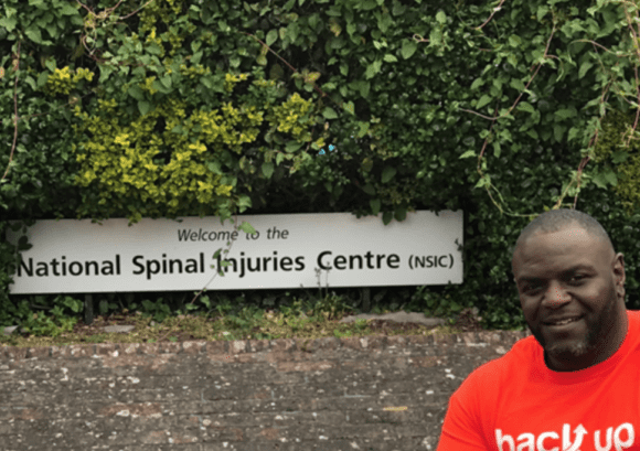 Shaun, one of our wheelchair skills trainers, posing with the sign outside the National Spinal Injuries Centre, Stoke Mandeville