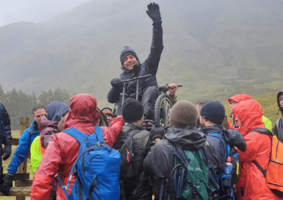 Andrew, a wheelchair user, being lifted over a fence during the Ben Nevis push