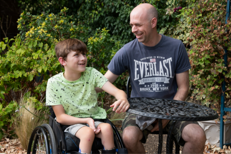 A wheelchair user child and his father in a garden. Support Back Up's service users by becoming a Back Up Spinal Cord Injury Mentor
