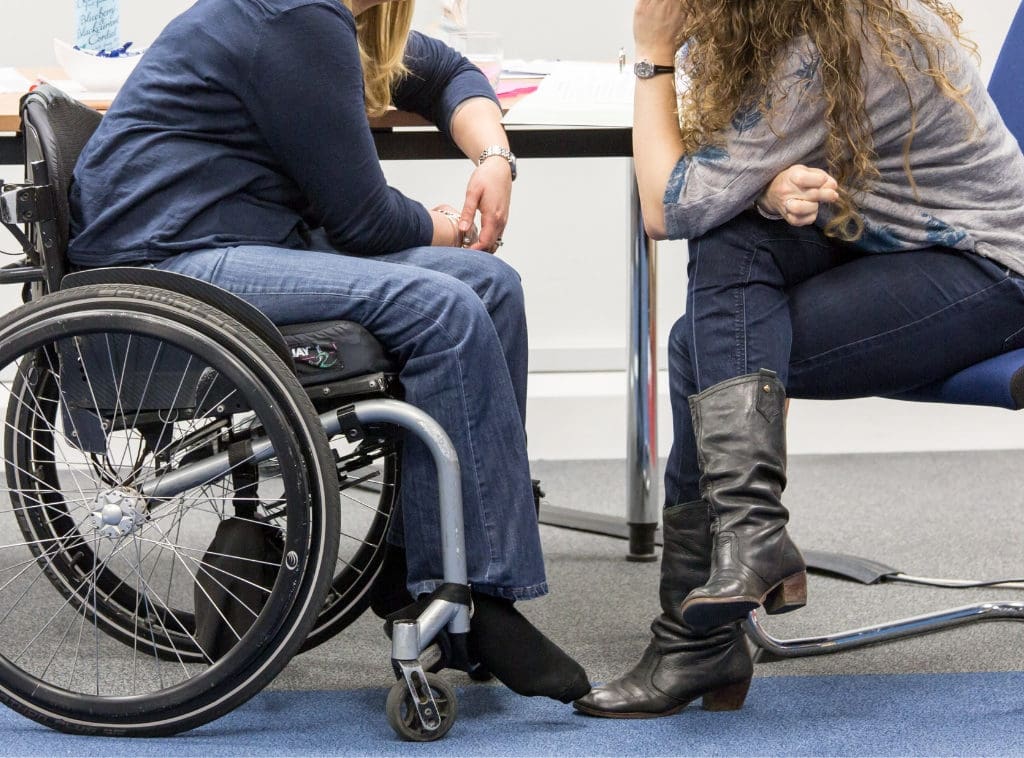 Two women talk by a table. One of them is a wheelchair user.