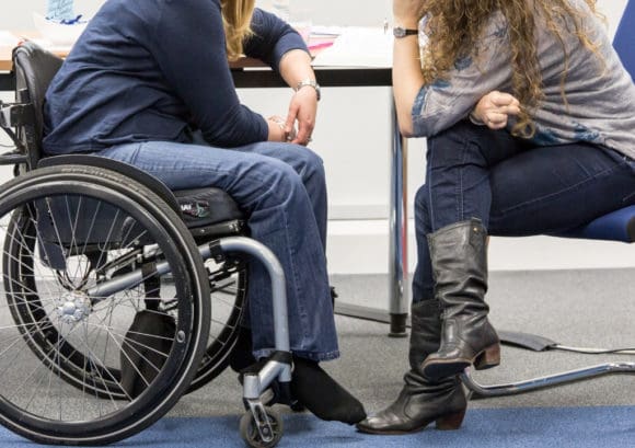 Two women talk by a table. One of them is a wheelchair user.