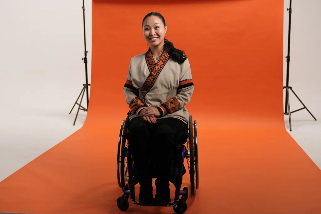 Limitless Ambition - the Back Up Strategy. Ayuna is on a film set with an orange background. She uses a manual wheelchair and is smiling at the camera.