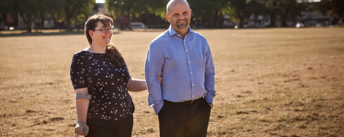 Chole and her partner are stood in a sunny field. She is using a crutch, both are smiling.