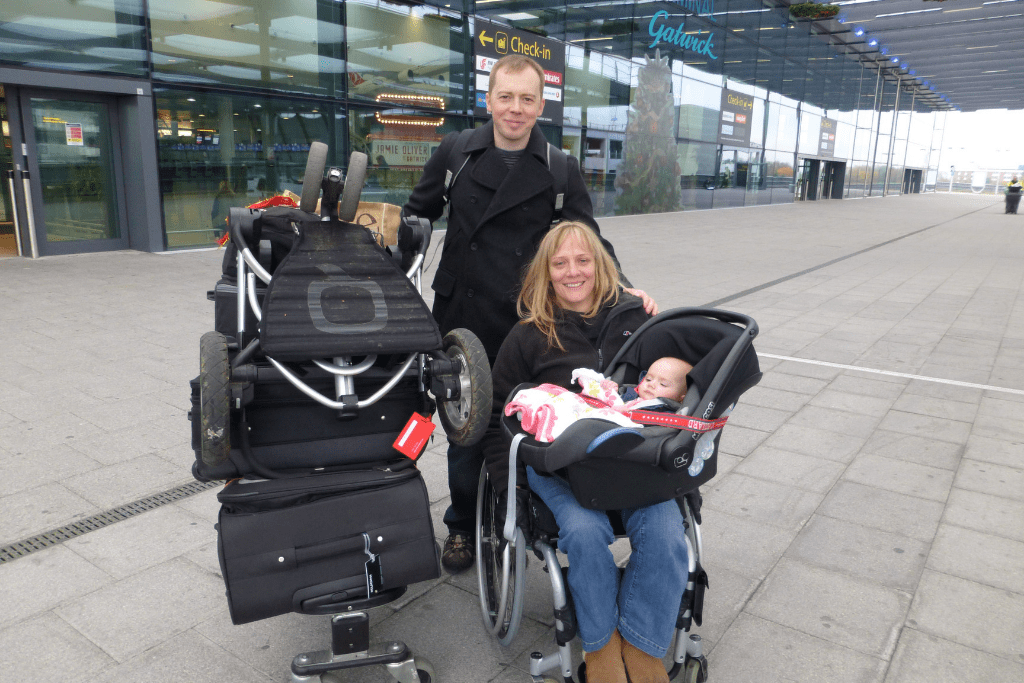 Clair in an airport with her luggage, her partner, and her baby daughter on her lap in a car seat