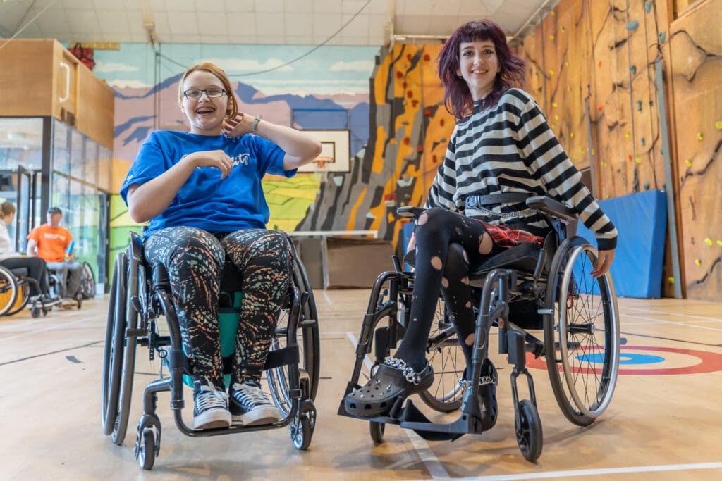 Two happy girls in their wheelchairs in a sports hall. 
