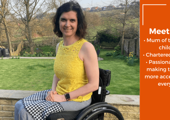 Katy is sitting in her wheelchair wearing a yellow t-shirt smiling in her garden. She has a C2-6 and T8 and T9 level incomplete spinal cord injury.